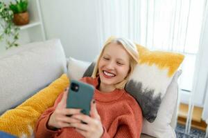 Young woman sit relax on sofa in living room browsing surfing wireless internet on smartphone, millennial girl rest on couch at home message text on modern cellphone, shopping online via website photo