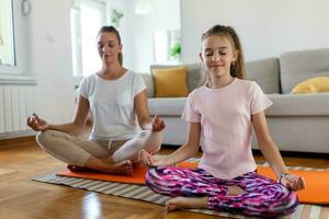 alegre joven mujer y pequeño niña en ropa de deporte hacer ejercicio juntos en moderno ligero habitación. madre y hija practicando yoga foto