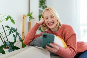 Attractive smiling woman using smart phone while sitting on the sofa at home. Communication and coziness concept. photo