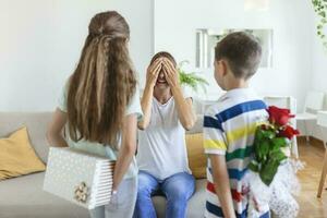 Cheerful little girl with gift box and youngest brother with bouquet of roses flowers smiling and congratulating happy mom on mother day at home. Happy Mothers Day photo