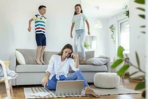 cansado joven madre sentado en el piso y trabajando con ordenador portátil y documentos mientras pequeño niños y saltando en el sofá y teniendo divertido y haciendo ruido foto