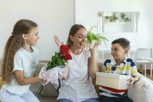 Cheerful little girl with bouquet of roses flowers and youngest brother with gift box smiling and congratulating happy mom on mother day at home. Happy Mothers Day photo