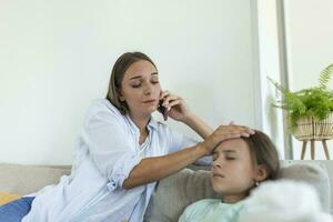 Mother measuring temperature of her ill kid. Sick child with high fever laying in bed and mother holding thermometer. Mother with cell phone calling to doctor photo