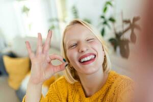Mischievous young woman takes selfie at home. Beautiful blond woman ok sign with hand blinking with one eye and with her tongue out photo