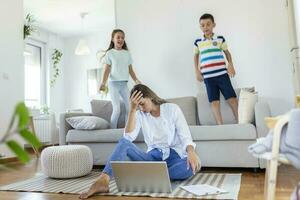 Tired young mother sitting on the floor and working with laptop and documents while little kids and jumping on the sofa and having fun and making noise photo