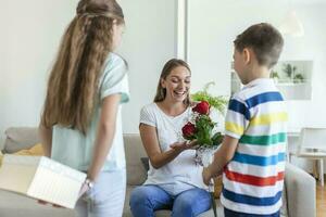 Happy children giving gift an flowers to mother. Happy Mothers Day Children boy and girl congratulate smiling mother, give her flowers bouquet of roses and a gift box during holiday celebration photo