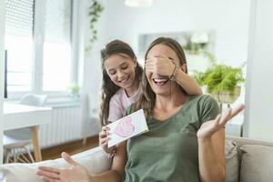 Happy little girl congratulating smiling mother and giving card with red heart during holiday celebration at home, covering her eyes and surprising her photo