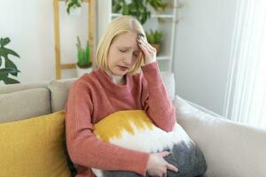 Portrait of a young blond woman sitting on the couch at home with a headache and migraine . Beautiful woman suffering from chronic daily headaches. Sad woman holding her head because sinus pain photo