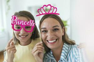 Funny family on a background of bright wall. Mother and her daughter girl with a paper accessories. Mom and child are holding paper crown on stick. photo