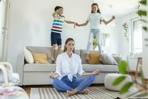 joven madre con cerrado ojos meditando en loto actitud en piso molesto a salvar interior armonía mientras emocionado niños saltando en sofá y gritando en ligero espacioso vivo roo foto