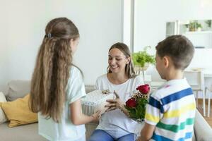 Happy children giving gift an flowers to mother. Happy Mothers Day. Children boy and girl congratulate smiling mother, give her flowers bouquet of roses and a gift box during holiday celebration photo