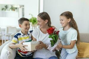 Happy children giving gift an flowers to mother. Happy Mothers Day. Children boy and girl congratulate smiling mother, give her flowers bouquet of roses and a gift box during holiday celebration photo