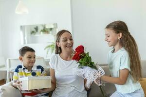 alegre pequeño niña con ramo de flores de rosas flores y el más joven hermano con regalo caja sonriente y felicitando contento mamá en madre día a hogar. contento madres día foto