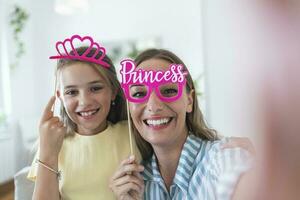 Funny family on a background of bright wall. Mother and her daughter girl with a paper accessories. Mom and child are holding paper crown on stick. photo