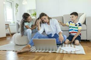 Small mischievous kids noising and distracting mother freelancer trying to concentrate on laptop and squeezing head working at table in light living room photo