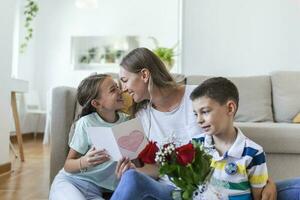 Young mother with a bouquet of roses laughs, hugging her son, and cheerful girl with a card and roses congratulates mom during holiday celebration at home. mothers day photo