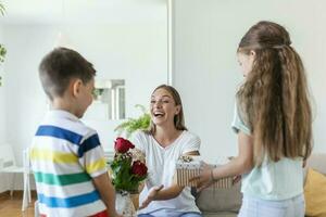 alegre pequeño niña con regalo caja y el más joven hermano con ramo de flores de rosas flores sonriente y felicitando contento mamá en madre día a hogar. contento madres día foto