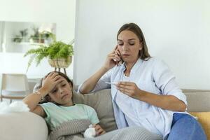 Mother measuring temperature of her ill kid. Sick child with high fever laying in bed and mother holding thermometer. Mother with cell phone calling to doctor photo