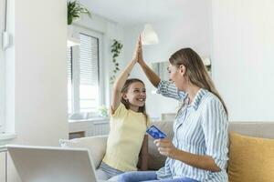 Low angle of excited small daughter giving high five to mother and screaming while celebrating successful online shopping using laptop at table in light living room photo