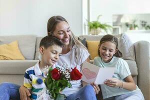 joven madre con un ramo de flores de rosas risas, abrazando su hijo, y alegre niña con un tarjeta y rosas felicita mamá durante fiesta foto