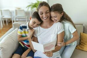 alegre joven madre y pequeño niños sentado en sofá y abrazando mientras leyendo deseos y Felicidades con fiesta en presentado tarjeta postal a hogar foto