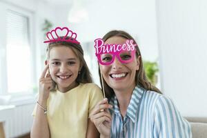 Funny family on a background of bright wall. Mother and her daughter girl with a paper accessories. Mom and child are holding paper crown on stick. photo