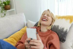 Young woman sit relax on sofa in living room browsing surfing wireless internet on smartphone, millennial girl rest on couch at home message text on modern cellphone, shopping online via website photo