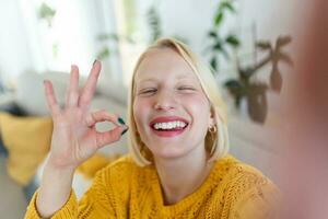 Mischievous young woman takes selfie at home. Beautiful blond woman ok sign with hand blinking with one eye and with her tongue out photo