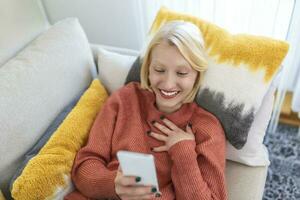 Young woman sit relax on sofa in living room browsing surfing wireless internet on smartphone, millennial girl rest on couch at home message text on modern cellphone, shopping online via website photo