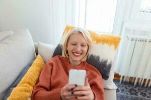 Young woman sit relax on sofa in living room browsing surfing wireless internet on smartphone, millennial girl rest on couch at home message text on modern cellphone, shopping online via website photo