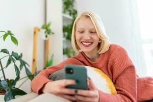 Young woman sit relax on sofa in living room browsing surfing wireless internet on smartphone, millennial girl rest on couch at home message text on modern cellphone, shopping online via website photo