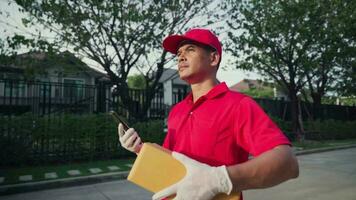 un entrega hombre vistiendo un rojo camisa camina alrededor participación un paquete o empaquetar caja y utilizando su teléfono a buscar para el clientes casa. video