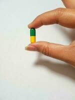 Isolated white photo of a hand holding a green and yellow medicine capsule.