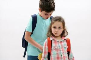 Mischievous kid girl makes faces at camera while her brother puts workbooks inside her backpack over white background photo