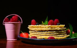 Still life. A plate of pancakes with fresh berries and mint leaves near a bucket with strawberries over dark background. photo