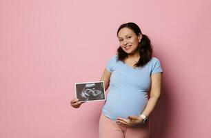 Gravid woman smiling at camera, holding baby ultrasound scan and caressing her pregnant belly, isolated pink background photo