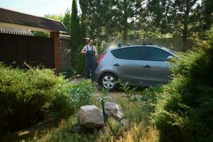 Young man in work overalls, using a water high pressure cleaners, washes his car in the backyard photo