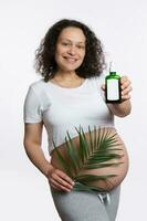 Smiling pregnant woman with palm leaf, shows at camera stretch marks massage oil, isolated on white. Skin and body care photo