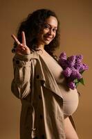 Fashion stylish pregnant woman in trench coat and beige lingerie, showing peace sign, holding a bunch of beautiful lilac photo