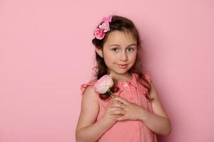 auténtico caucásico hermosa elegante pequeño niña en rosado vestido, participación un Rosa flor, sonriente lindo mirando a cámara foto