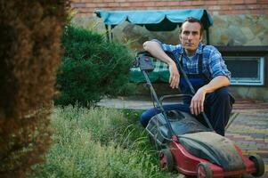 profesional masculino jardinero en jardinería uniforme, sentado cerca eléctrico césped cortacésped, sonriente mirando con confianza a cámara foto