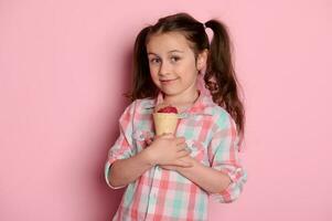 Lovely kid girl holds a waffle cone with chocolate strawberry ice cream, smiles looking at camera, isolated on pink photo