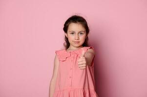 Cute little girl in elegant pink dress, gestures with thumb up, expressing positive emotions, smiles looking at camera photo