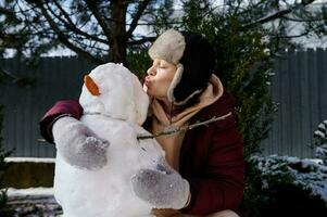 Pretty woman in warm ear flaps hat, hugging and kissing a snowman, enjoying winter leisure games at Christmas holidays photo