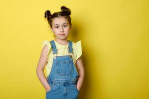 Beautiful smart little kid, school girl in yellow t-shirt, holding hands in pockets of jeans overalls, looking at camera photo