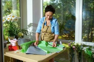 Beautiful woman florist laying color wrapping paper on the table while arranging flower bouquet in floral design studio photo