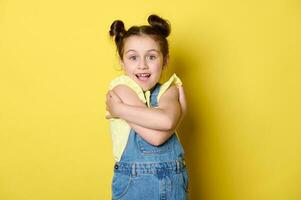 Happy emotional little kid girl embraces herself, smiles at camera, expressing positive emotions over yellow background photo
