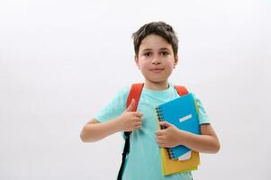 inteligente colegio niño, hermoso preadolescente chico sonrisas mirando a cámara, pulgar arriba, demostración aprobación firmar en blanco fondo foto