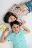 Top view adorable kids, mischievous sister and brother eating pop corn looking at camera, lying over white background. photo