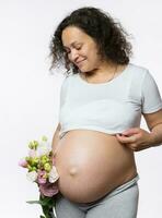 Beautiful multi ethnic gravid woman holding bunch of flowers, smiling looking at her pregnant belly in late pregnancy photo